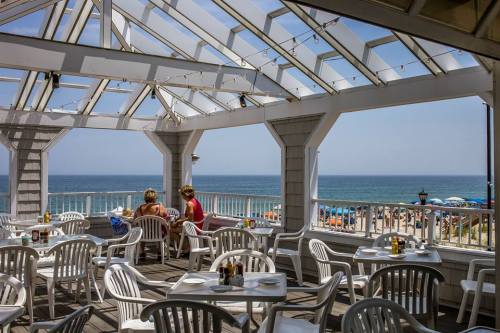 A beachside restaurant with white chairs and tables, overlooking the ocean and colorful umbrellas on the sandy shore.