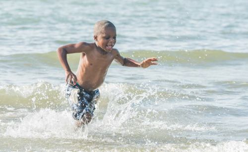 A young boy runs joyfully through shallow water at the beach, splashing as he plays in the waves.