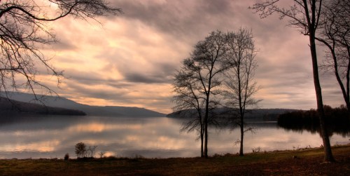 A serene lake at sunset, surrounded by bare trees and soft clouds reflecting on the water's surface.