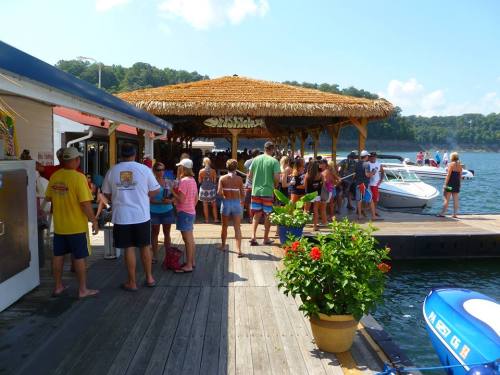 A lively waterfront scene with people gathered near a tiki bar, boats docked, and lush greenery in the background.