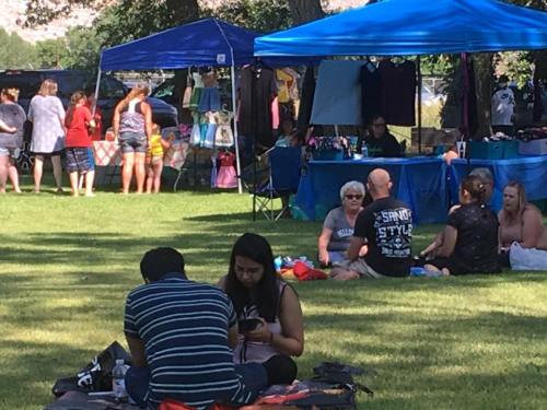 A busy outdoor market scene with people shopping and relaxing on the grass under blue tents.