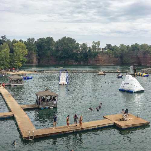 A lake with people swimming and playing on inflatable structures, surrounded by trees and a wooden dock.