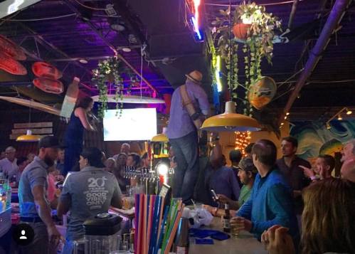 A lively bar scene with people enjoying drinks, a performer on a table, and colorful decorations overhead.