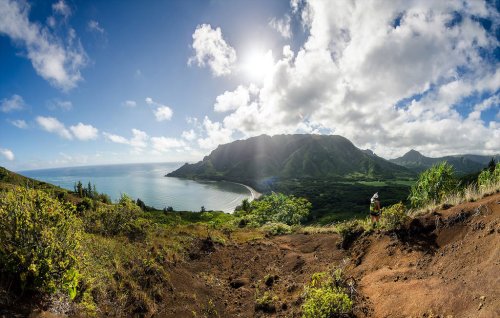A panoramic view of a lush landscape with mountains, a coastline, and a bright sky with scattered clouds.