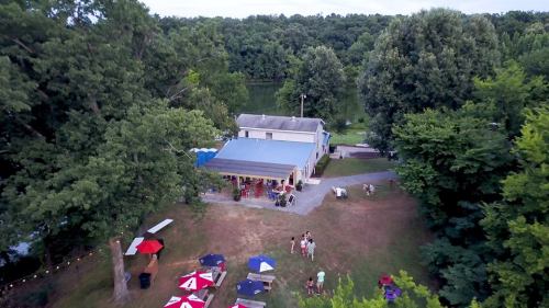 Aerial view of a gathering area with a house, trees, and people enjoying a sunny day by a river.