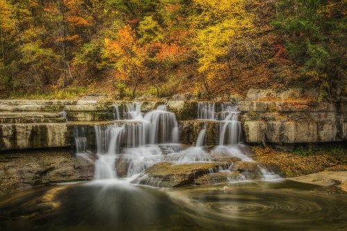 A serene waterfall cascades over rocks, surrounded by vibrant autumn foliage in warm hues of orange and yellow.