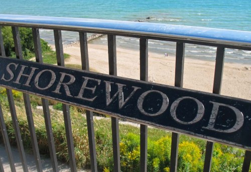 Sign reading "Shorewood" in front of a beach and blue water, with greenery in the foreground.