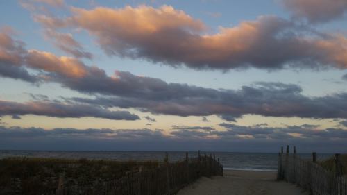 A sandy path leads to the beach, framed by a wooden fence under a sky filled with clouds at sunset.