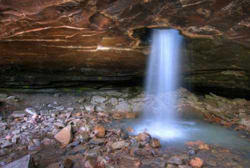 A serene waterfall cascades from a rocky overhang into a pool, surrounded by stones in a cave-like setting.