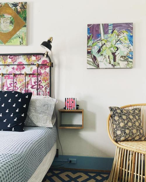 A cozy bedroom featuring a patterned bed, decorative pillows, a wall painting, and a rattan chair.