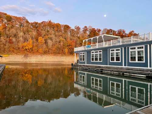 A lakeside building reflects on calm water, surrounded by autumn foliage and a clear sky with a visible moon.