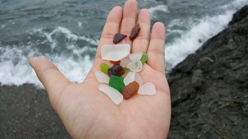A hand holding various pieces of colorful sea glass against a backdrop of waves and rocky shore.
