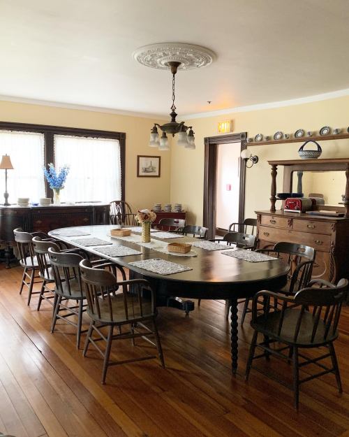 A spacious dining room with a large wooden table, surrounded by chairs, and warm wooden floors. Natural light filters in.