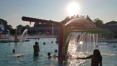 Children and adults play in a sunny pool with colorful water features and fountains, creating a fun summer atmosphere.