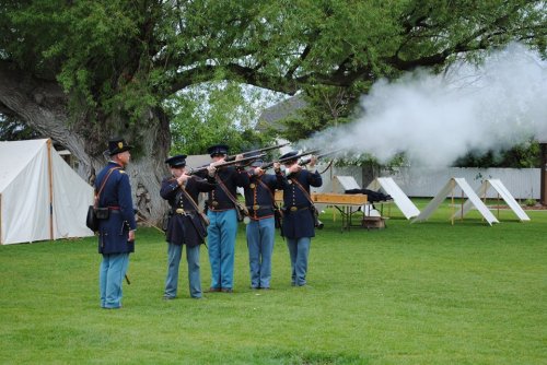 A group of soldiers in historical uniforms firing rifles, with tents and trees in the background. Smoke billows from the guns.