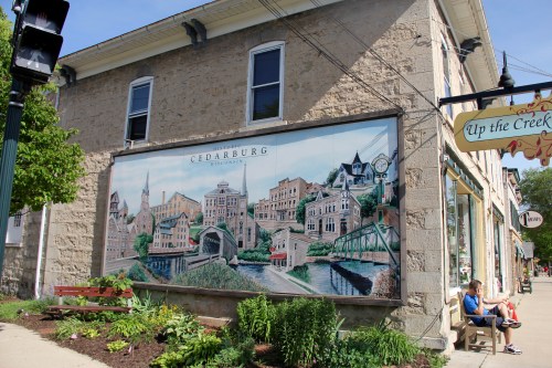 A mural depicting a scenic view of Cedarburg, Wisconsin, on the side of a stone building, with a bench nearby.