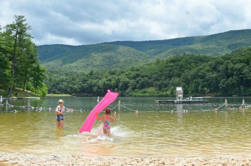 A girl splashes in the water with a pink float, while an adult watches nearby on a sandy beach by a lake.