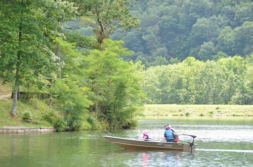 A small boat with two people glides across a calm lake surrounded by lush green trees and hills.