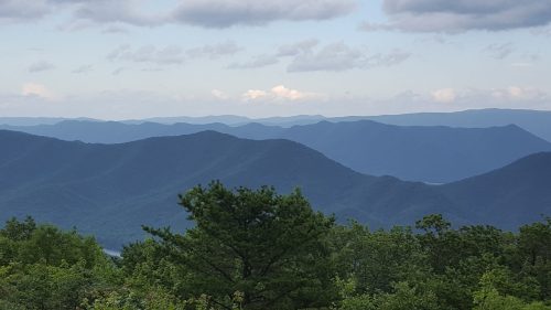 A panoramic view of layered blue mountains under a cloudy sky, with green trees in the foreground.