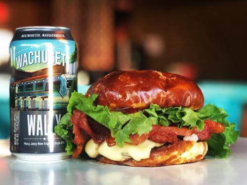 A pretzel bun burger with lettuce, tomato, and sauce next to a can of Wachusett beer on a table.