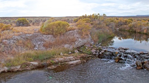 A serene river flows through a landscape of rocky banks and autumn-colored vegetation under a cloudy sky.