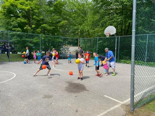 A group of children playing with colorful balls on an outdoor basketball court surrounded by trees.