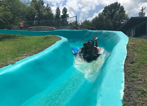 A person slides down a bright blue water slide, splashing into the water below on a sunny day.