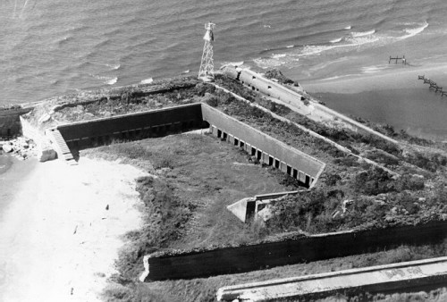 Aerial view of a coastal fortification with a lighthouse, surrounded by water and overgrown vegetation.