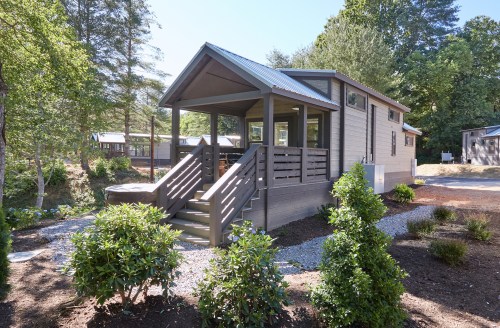 A modern cabin with a porch, surrounded by greenery and gravel pathways, set in a wooded area.