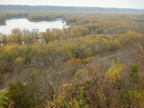 A scenic view of a river winding through a forest with autumn foliage and distant hills under a cloudy sky.