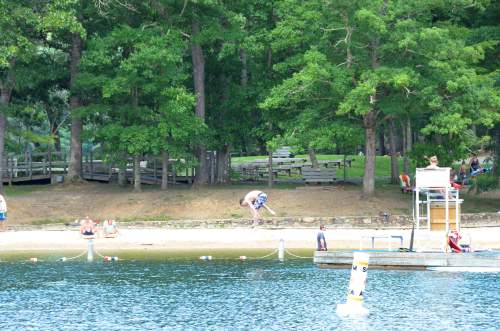 A serene lakeside scene with people relaxing on the beach, trees in the background, and a lifeguard station nearby.