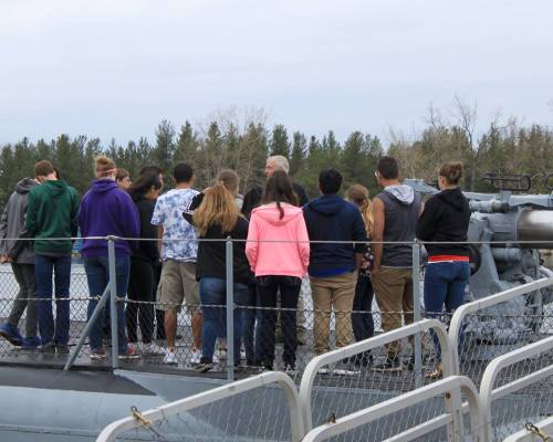 A group of people stands on a boat, facing away, with trees and water in the background.