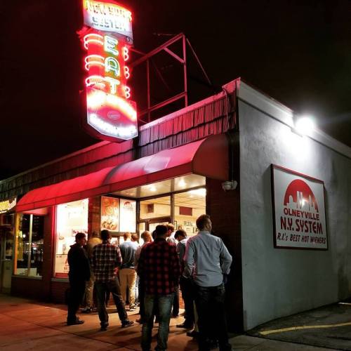 A group of people stands outside a diner with neon signs, waiting to enter the Olneyville N.Y. System restaurant at night.