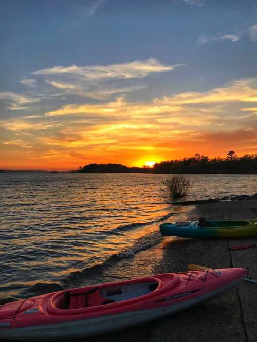 A serene sunset over a lake, with colorful kayaks resting on the shore and vibrant clouds in the sky.