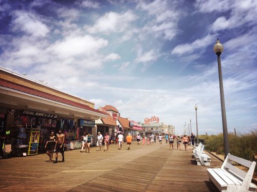 A bustling boardwalk with shops, people walking, and a clear blue sky overhead. Benches line the wooden path.