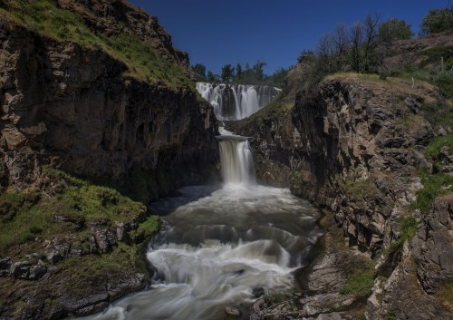 A scenic waterfall cascading down rocky cliffs into a river, surrounded by lush greenery under a clear blue sky.