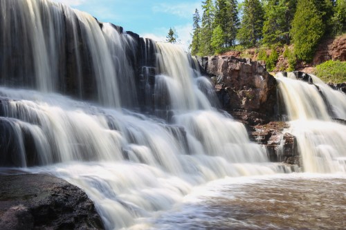 A cascading waterfall flows over rocky cliffs, surrounded by lush green trees under a clear blue sky.