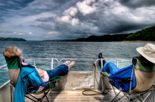 Two people relax in chairs on a boat, with their feet up, overlooking a calm lake under a cloudy sky.