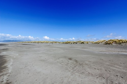 A wide sandy beach under a clear blue sky, with grassy dunes in the background and scattered clouds.