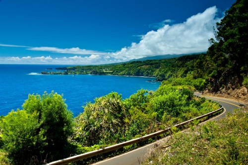 Scenic coastal view with a winding road, lush greenery, and vibrant blue ocean under a clear sky with clouds.
