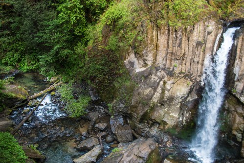 A serene waterfall cascades over rocky cliffs, surrounded by lush greenery and a flowing stream below.