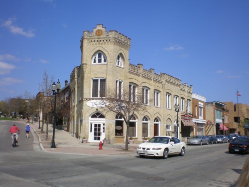Historic stone building with arched windows, surrounded by trees and parked cars, on a sunny street.