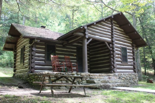 A rustic log cabin surrounded by trees, featuring a porch with rocking chairs and a picnic table in front.