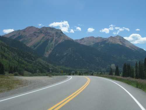 A winding road leads through lush green hills and mountains under a clear blue sky with scattered clouds.