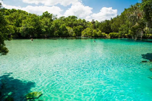 A serene, turquoise lake surrounded by lush greenery and a clear blue sky, with a few people swimming.
