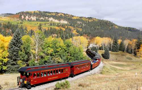 A red train winds through a scenic landscape of autumn trees and mountains under a cloudy sky.