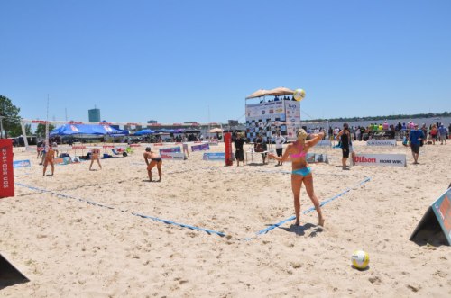 A beach volleyball match in progress, with players on sand and spectators in the background under a clear blue sky.