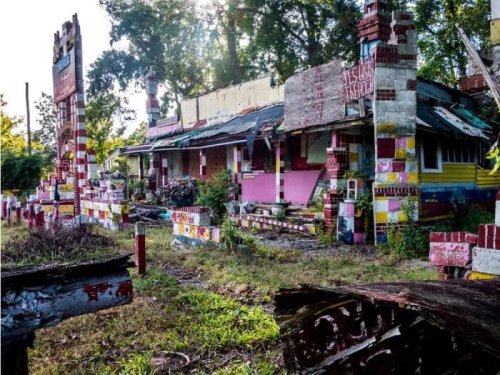 Abandoned colorful building with graffiti, surrounded by overgrown grass and trees, under a bright sun.