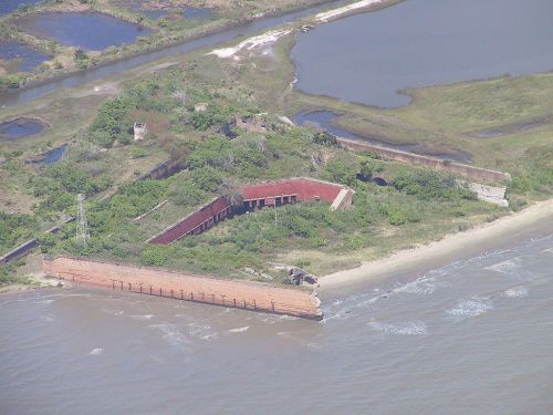 Aerial view of a coastal area featuring abandoned structures, overgrown vegetation, and a sandy shoreline.
