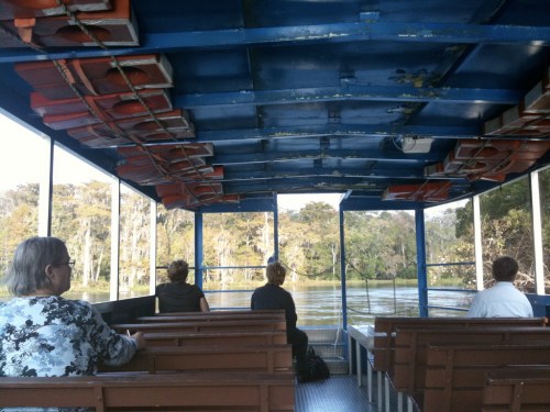 Interior of a boat with passengers seated, looking out at a scenic view of trees and water. Life jackets are visible above.